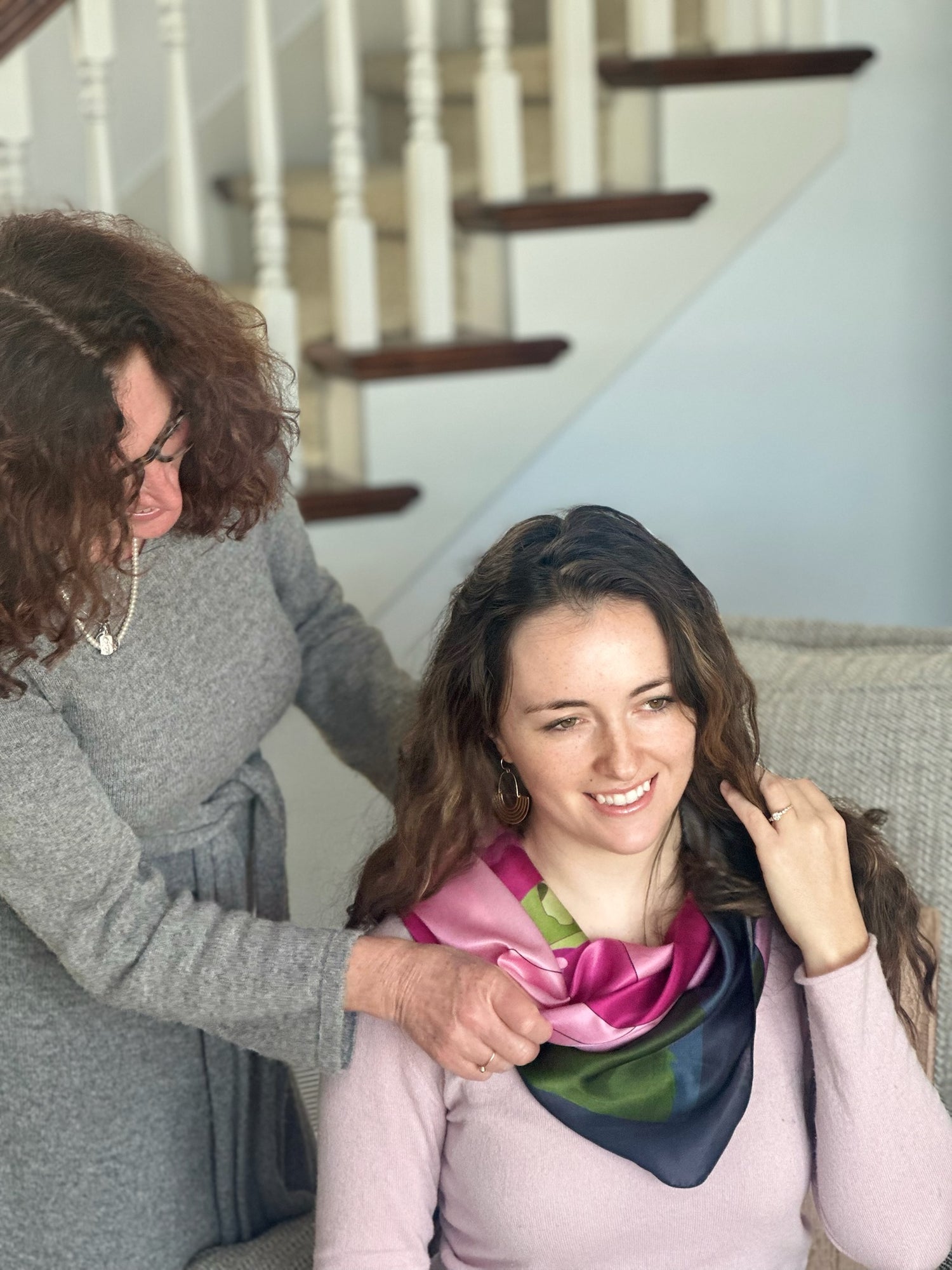 Woman tying the green and pink silk, Garden of Impossible scarf on daughters's neck in a home setting.