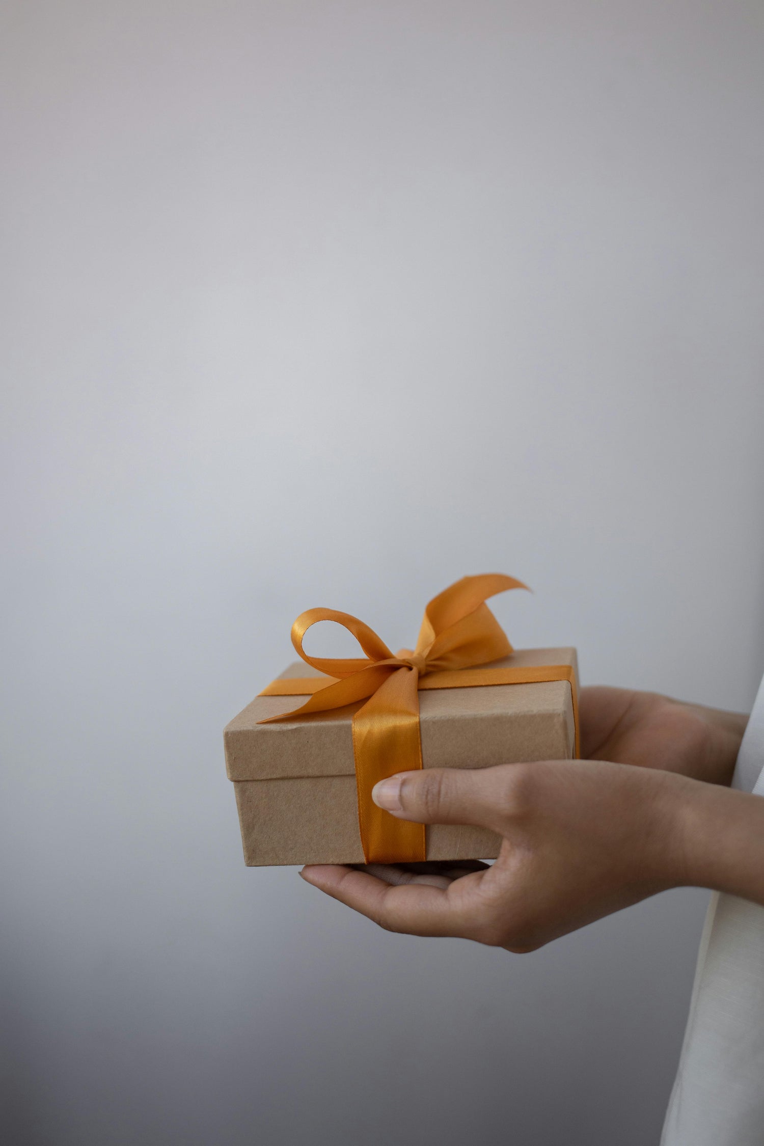 Hand holding a small brown gift box with an orange ribbon against a plain background